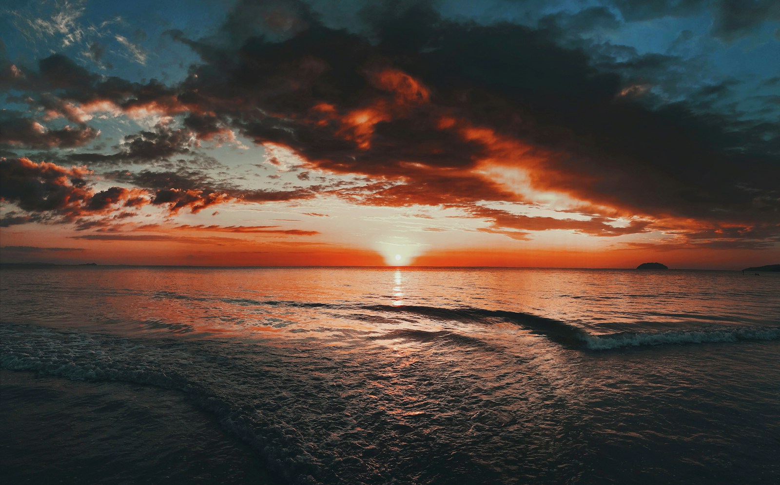 丹绒亚路海滩的橙红晚霞铺满海平线，落日与浪花在海面映出金色光影